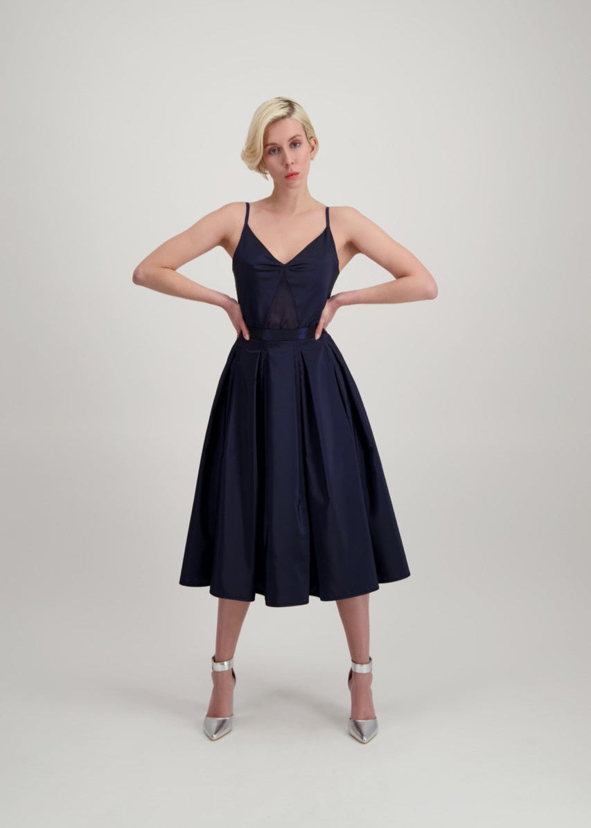 Femme blonde, les mains à la taille, habillée d'un débardeur et d'une jupe bleu marine.