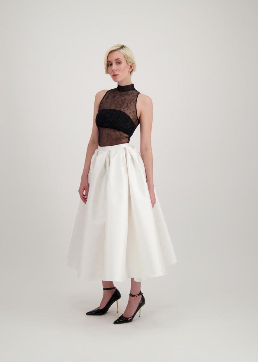 Femme blonde de profil habillée d'une jupe midi blanche et d'un body transparent noir laissant apparaître un bandeau noir