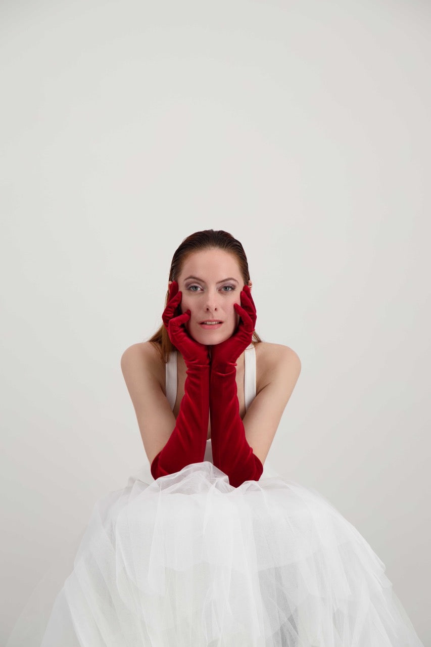 Jeune femme rousse qui se tient le visage avec ses mains. Elle porte une robe blanche et des longs gants en velours rouges