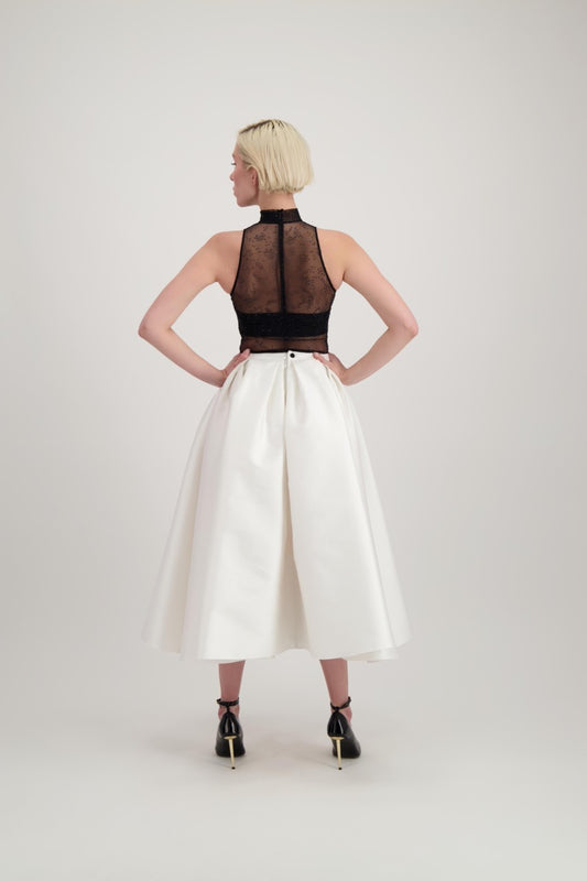 Femme blonde de dos habillée d'une jupe blanche midi et d'un body transparent noir laissant apparaître un bandeau noir