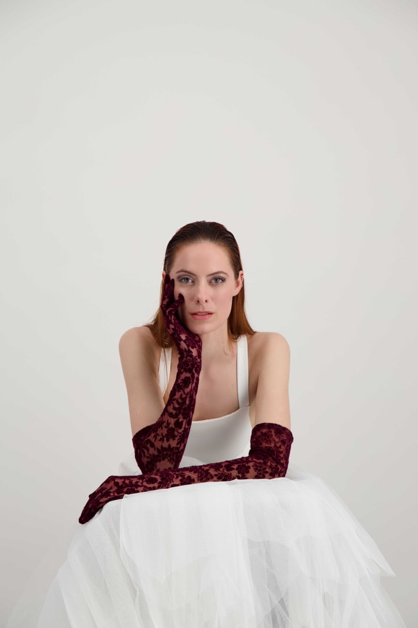 Jeune femme rousse qui tient la tête avec sa main. Elle porte une robe blanche et des longs gants bordeaux aux motifs fleuris