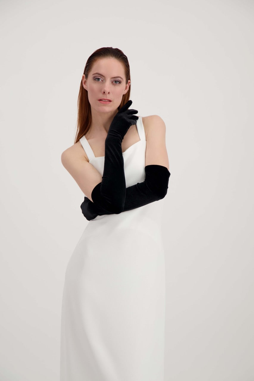 Jeune femme rousse, les bras croisés et une main sous le visage. Elle porte une robe blanche et des longs gants noirs en velours