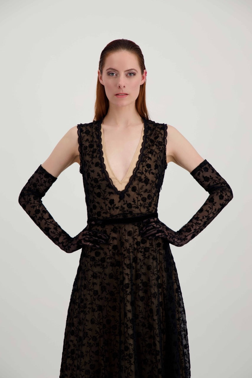 Jeune femme rousse qui a les mains sur les hanches. Elle est habillée avec une robe en dentelle noire et des longs gants assortis